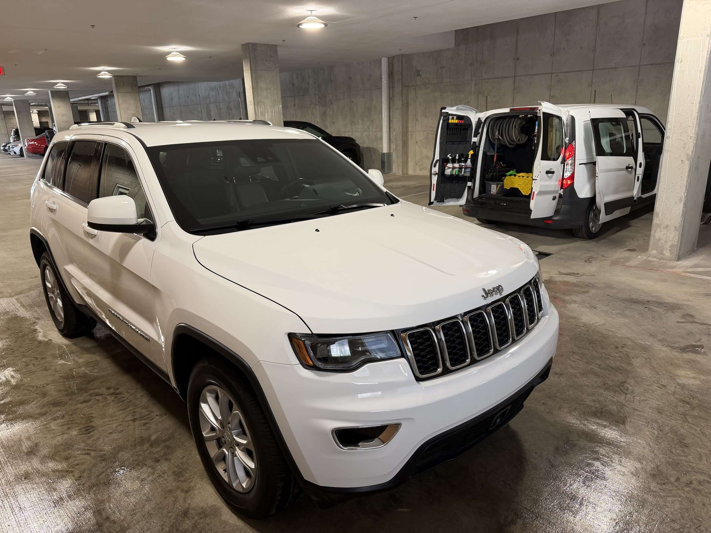 Freshly detailed white Jeep Grand Cherokee in a parking garage, with a mobile detailing van open behind it showing its onboard equipment